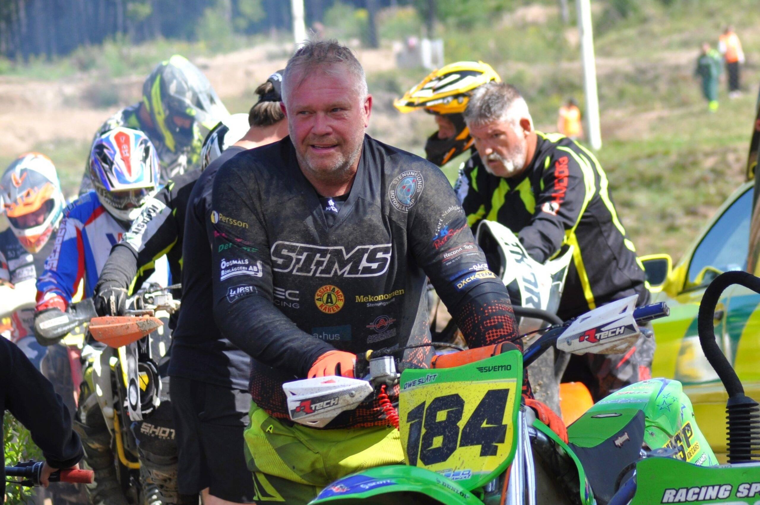 En bild på crossförare Owe Slätt som står bredvid sin hoj med crossutrustning men utan hjälm och ser glas och svettig ut. I bakgrunden syns andra crossförare.